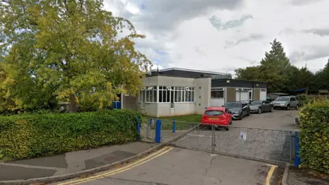 Google A grey building behind blue gates. It is off a leafy road and cars and parked outside. 