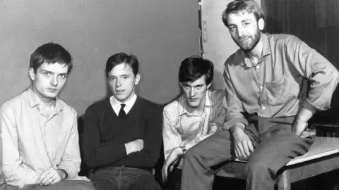 Black-and-white image of Joy Division. From left: Ian Curtis, with short dark hair and an open-necked shirt, Bernard Sumne, in a dark sweater, thin black tie, Stephen Morris with short dark hair and an open-necked shirt, and Peter Hook, with open-necked shirt - he has lighter hair and a beard. He is sitting on a table a little in front of the others.