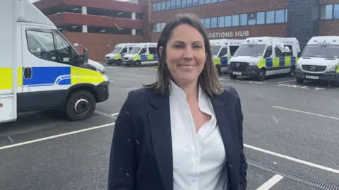 Sparks is stood in the car park of a police station with police vans parked up behind her. She has a white shirt and blue jacket on.