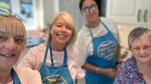 FODK A group of four women looking at the camera and smiling. Three are wearing blue aprons with yellow writing on. Jan Kellond, on the right of the picture, is wearing a flowery top. 