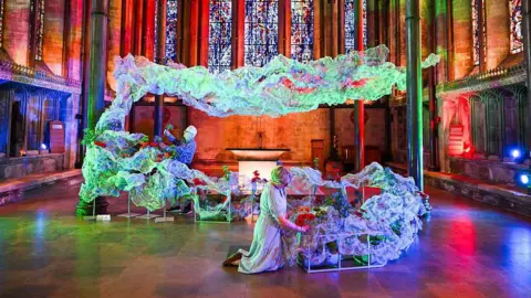 Finnbarr Webster/Getty Images A floral display with lots of material shaped to look as though it is floating in a cathedral with dramatic coloured lighting. Two people making finishing touches with flowers.