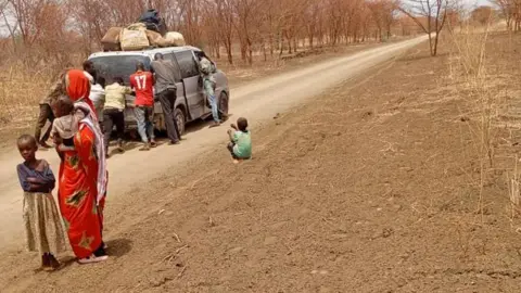 Amira Men pushing a vehicle loaded with baggage on its roof along a dirt road in Sudan.