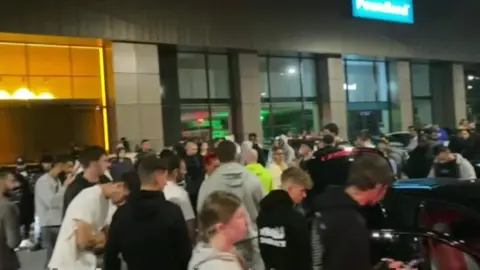 BBC Image showing crowds of people milling between cars at a retail park in Enfield. A Poundland store can be seen in the background