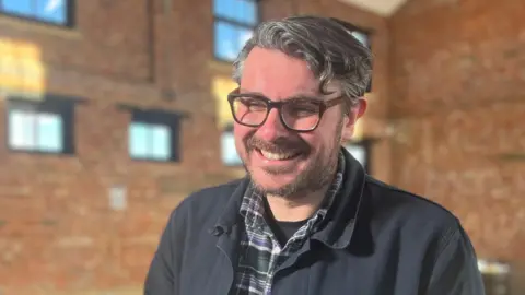 Ross Millard, smiling at the camera. He is wearing a green and purple checked shirt and a dark blue jacket. He has short wavy hair and glasses. 