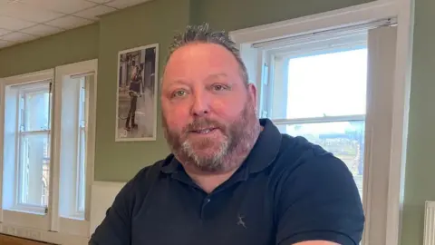 Charles Heslett/BBC A man sits at a table in a room with green walls and large white windows. He is looking at the camera. He has short grey hair and beard. He is wearing a navy blue polo shirt.