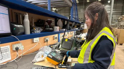 A woman with long brown hair, long sleeves and gloves looking at a piece of electronic equipment with keys and a display screen on a black worktop in a room with blue shelves behind the desk