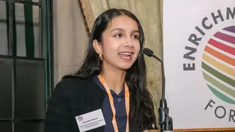 Duke of Edinburgh A woman talking into a microphone at a podium with a lanyard round her neck and an ID on her lapel. There is a large printed out crest or emblem to her left, which depicts the circle, or globe, shaded with various colours.
