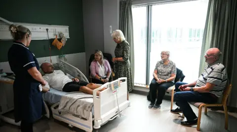 PA media queen Camilla stands next to the hospital bed and speaks to Paul Holdway, who is surrounded by family members