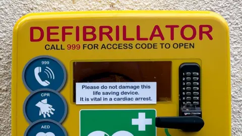 Yellow defibrillator box attached to a wall. It has a code lock and reads "call 999 for access code to open".