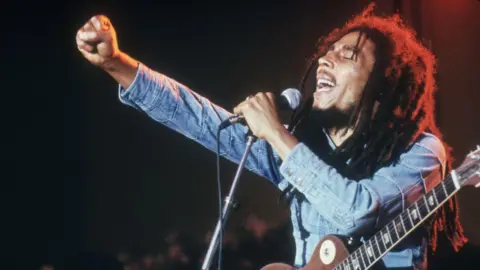 Getty Images Bob Marley signing in to a microphone, with his eyes closed. One hand is holding the microphone and then other has making a fist and is out in font of him. He is wearing and denim shirt and has a guitar over his shoulder.