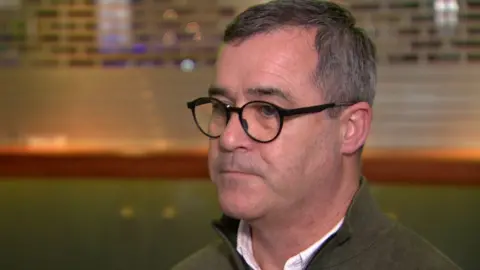 Alan Brecknell is being interviewed. He is standing slightly diagonal looking off camera. He has short, greying hair with circular dark framed glasses, a green zip up jumper and a white shirt. He has a neutral expression on his face.