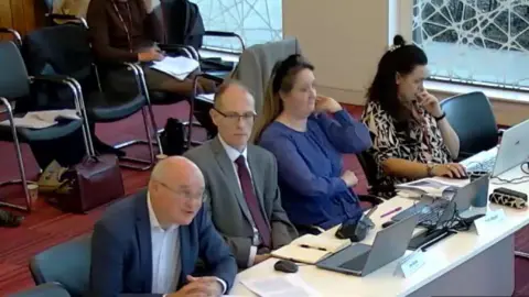 Worcestershire County Council A camera screenshot shows Simon Geraghty speaking at a council meeting at Wyre Forest House. He is wearing a white shirt and blue suit, and is sat on a table next to officers from Worcestershire County Council.