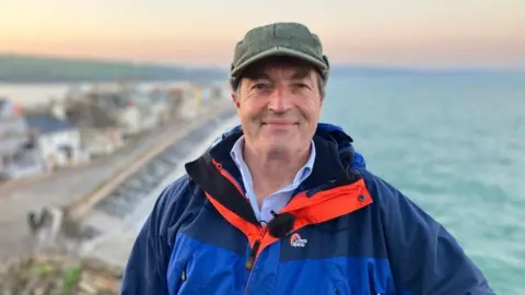 A picture of David Braine wearing a blue jacket and a green cap. He is stood in front of the sea and the seafront.