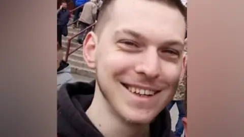 A head and shoulders shot of a man smiling. He has short dark hair and a dark hoodie