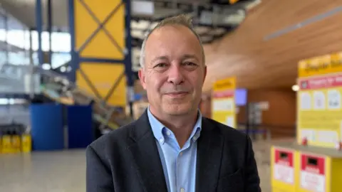 Nico Bezuidenhout, a man with short grey hair, wearing a light blue shirt and a dark blazer. He is smiling directly at the camera, while standing in the departures hall at Guernsey Airport.
