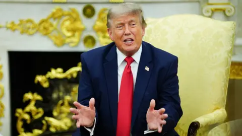 Donald Trump sits in the Oval Office wearing a navy blue suit and red tie.