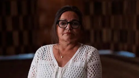 Gavin Hopkins A photo of Fee against a dark background. She has dark hair and glasses. She is wearing a white crocheted cardigan