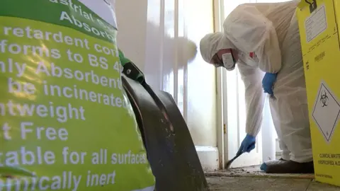 One of Jim Gildea's colleagues is bent over, wearing a white protective suit, face mask, gloves, shoe coverings and glasses as he scrapes grime from the floor. There is a bag of absorbent granules, a shovel and a burn box in the foreground.