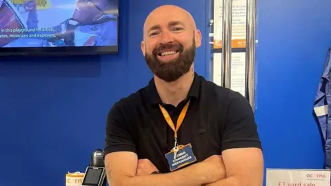 A man with a shaved head and a lustrous dark beard stands in front of a shop counter with a white top and a blue frontage, labelled "acorns". He is wearing a black polo shirt and black jeans and has his arms crossed in front of his body. He is smiling at the camera.
