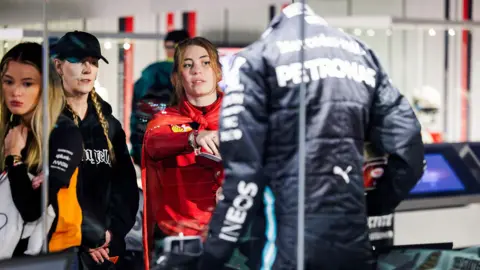 Three young women are looking at a Mercedes racing drivers suit in a cabinet at Silverstone Museum. one of the women, wearing a red top, is pointing at the suit. Another woman in a black top and cap is looking at the suit. A third young woman appears to be looking at another exhibit.