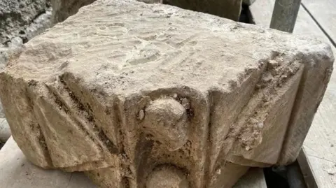 A large stone with carvings, including a triangle shape and a couple of sphere shapes. Sitting on a scaffolding board.