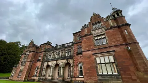 An imposing red sandstone building with stone archways and ornate features. There are decorative designs above the windows and a on the upper sections of the building's facade.