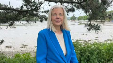 A woman with white blonde shoulder length hair wearing a blue jacket and cream t-shirt, standing on a river edge