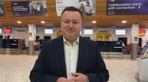 A man in a dark suit and white shirt with an open collar standing in Guernsey Airport check-in area. 