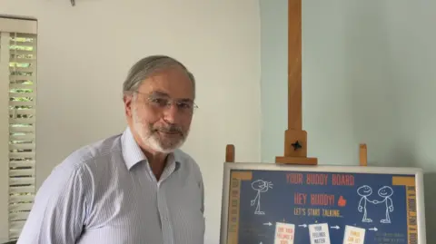 Morgan Webb/BBC Gentleman with a white and light blue striped business shirt on, glasses and a grey beard and hair, next to a "Buddy Board" for his charity Joe's Buddy Line.