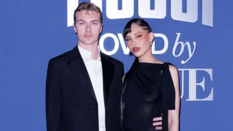 Getty Images Married couple Lucky Blue Smith and Nara Smith stand against a blue backdrop in formal wear. Nara is wearing a black dress with strands on each side and Lucky wers a white shirt and black blazer.
