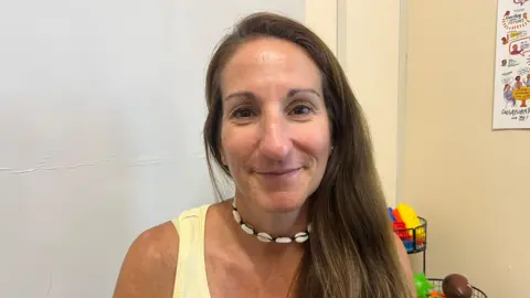 Kim is smiling at the camera. She is wearing a yellow top, a shell necklace and has long brown hair