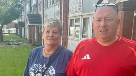 A man and a woman stand in front of a pile of rubbish on a front lawn by block of flats.