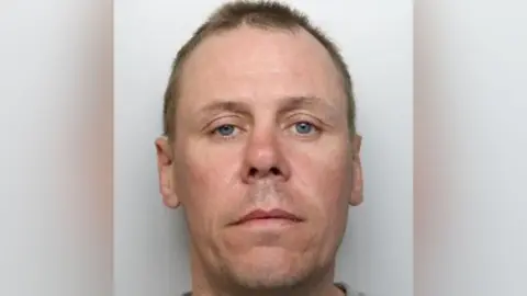 Mugshot of a man with short brown hair and an oval face. He has blue eyes and a slightly crooked nose. He has a neutral expression.