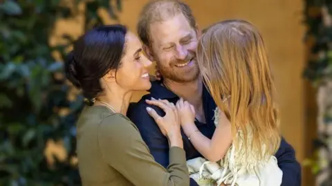 Instagram/Meghan, Duchess of Sussex Duke and Duchess of Sussex in casual outfits embracing and holding Lilibet in a summer dress, she has bare feet and her back is to the camera; she has long, red hair