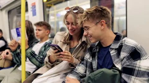 Getty Images A young adult male and a young adult female sit next to each other on a Tube looking at a mobile phone and smiling   