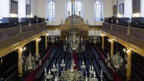 Getty Images High-angle view of the interior of Bevis Marks Synagogue, showing rows of dark wooden pews, an upper gallery balcony, and large, ornate brass chandeliers hanging from the ceiling facing a gilded Ark.