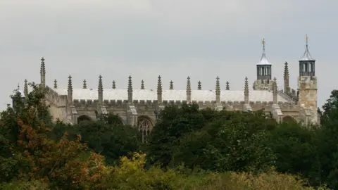 The chapel of Eton College, seen from the Thames at Windsor, Berkshire. It has a giant roof with two spires at one side. It pokes out of greenery. 