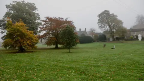 The misty Romaldkirk Village Green. There are several mature trees with yellow autumn leaves on the grass. A two-storey stone house is in the background. 