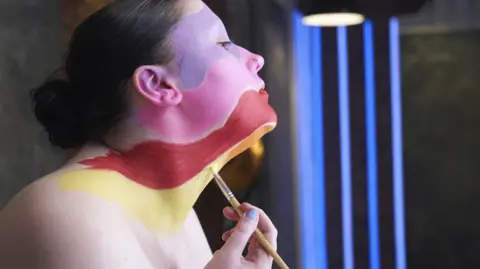 BBC/Wall To Wall/Dave King Ailish is in the middle of applying a make-up look to her face. We can see her from a side-on angle. Her head is tilted upwards and she is looking straight ahead, presumably into a mirror. She has a paintbrush in hand and is painting on a yellow colour underneath her chin. Ailish's face has been sectioned off into different colour blocks with gaps of unpainted skin in between. The upper part of her cheek and one of the eyes is lilac. The middle of her face and nose is pink. Her lower lip and upper chin is red. Part of her lower chin is orange and underneath that is the yellow on her neck. Ailish's black hair has been scraped back into a low bun and she has a silver hoop nose piercing. The background is out of focus but she is in a dark room with neon blue vertical lines of light.