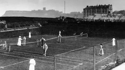 Hulton Archive via Getty Images People playing tennis at Scarborough in about 1913. The Floral Hall, a large glass building is behind the tennis courts.