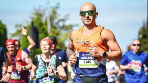 Adriano Picano A man wearing a running vest and sunglasses running in a crowd of others. He has tattoos on one arm and the sky is blue.