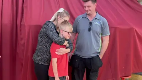 Ryan standing in a red t-shirt and glasses, with his mum giving him a hug. The pair are also standing next to Ryan's dad, who is wearing a grey t-shirt and black trousers