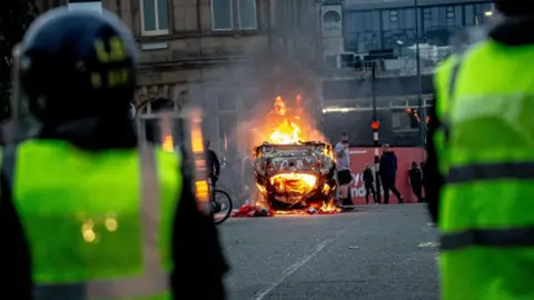 Getty Images Taken from behind two police in riot gear, a car burns