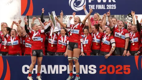 Getty Images A group of women rugby players celebrating and jumping in the air holding a trophy.