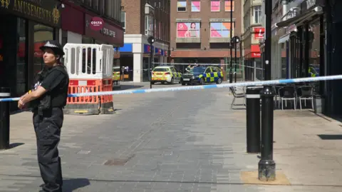 Ant Saddington/BBC A police officer in a black uniform stands by a blue and white police cordon in front of a wide alleyway. Two police cars are parked at the other end of the road.