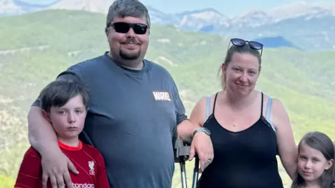 A red liverpool FC football shirt handout a boy with a neutral expression, standing next to a man in sunglasses with a gray Marvel T-shirt. Smiling, there is one hand around your son and the other one is resting on a pair of crutches. She is next to a woman with her hair and has sunglasses on her head, smiling from her arm and a short girl with brown hair. They stand in front of the mountains in Türkiye.