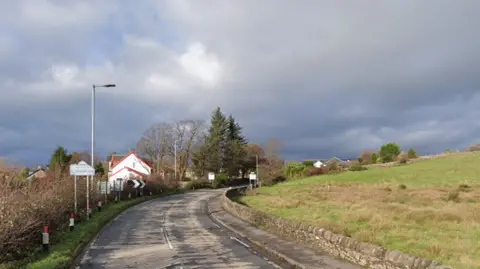 A stretch of road, entering a rural town. A sign saying Welcome to Cardross is visible.