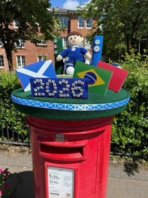 Bill McGowan A postbox with a knitted Scotland football player sitting on, with mocked up envelopes showing the flags of Scotland, Brazil and Morocco. A knitted 2026 sign sits in front on them, still on the top of the post box.