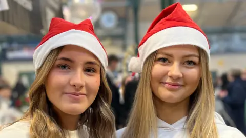 Two girls with long blonde hair, they are wearing Santa hats and white jumpers