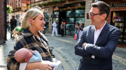 PA Media Prime Minister Andy Burnham speaks with a woman in Belfast, as he makes his first visit to Northern Ireland since coming to office. He has dark hair, wearing glasses, a navy blazer and white shirt. His arms are folded. The woman has long blonde hair tied back, wearing a check coat and carrying a baby.